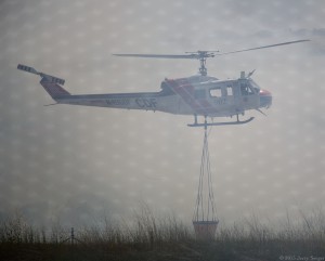The helicopter rises from loading at the pond, shrouded by smoke.